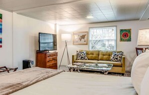 Habitación de hotel con cama, sofá, mesita de centro, televisión, cómoda y ventana con persianas.