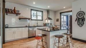 A modern kitchen with white cabinetry, a central island with stools, a built-in oven, and a window providing natural light. - The Sage Estate - Only 6 Minutes from Downtown (Nashville)