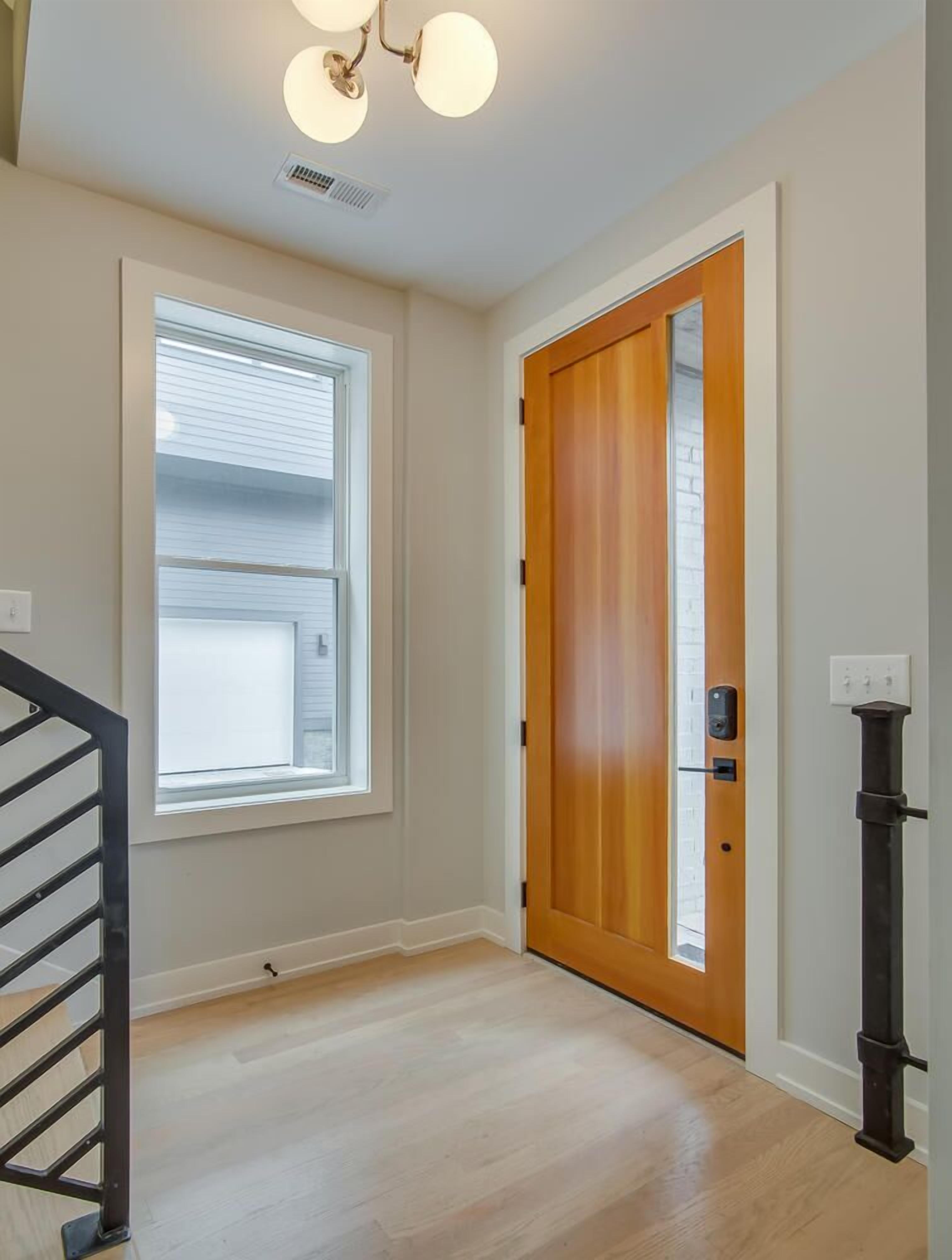 A hallway with a wooden door, a window with blinds, a light fixture, and a black railing.