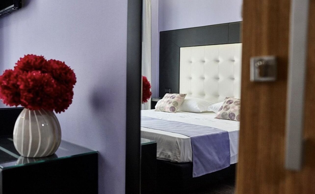 Una camera d'albergo con un letto, un comodino e un vaso di fiori rossi.