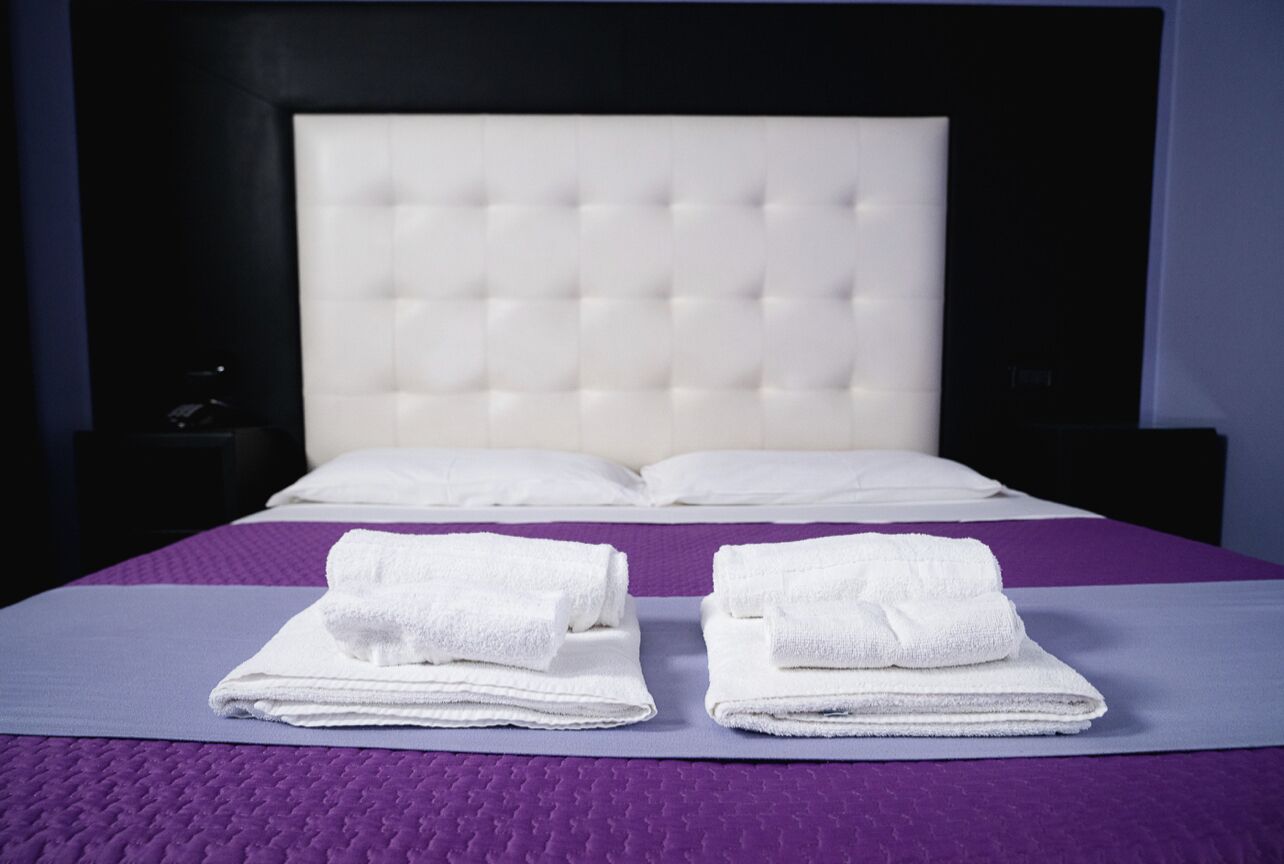 A neatly made bed with white linens and a purple bedspread, a headboard, and a small bedside table with a lamp.