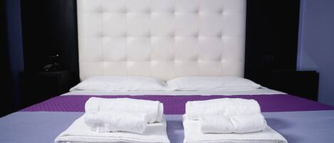 A neatly made bed with white linens and a purple bedspread, a headboard, and a small bedside table with a lamp.