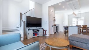 A modern living room with a flat-screen TV, a staircase, a kitchen area, and a dining space.