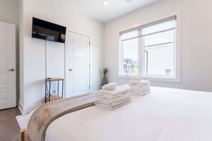 Habitación de hotel con cama, televisor montado en la pared, ventana con vista a edificios y una mesita con una planta.