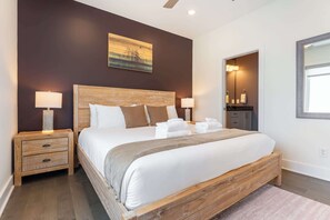 A bedroom with a wooden bed, two bedside tables with lamps, a painting on the wall, and a bathroom visible through an open door. - The East Side Vibe- Modern East Nash Stay, Garage & Open Living + Great Location (Nashville)