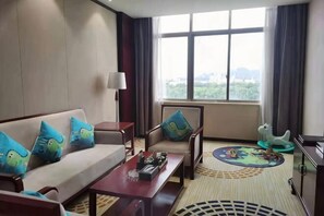 Habitación de hotel con sofá, sillones, mesa de centro, lámpara, vista a la ciudad y una pequeña mesa con un objeto decorativo.