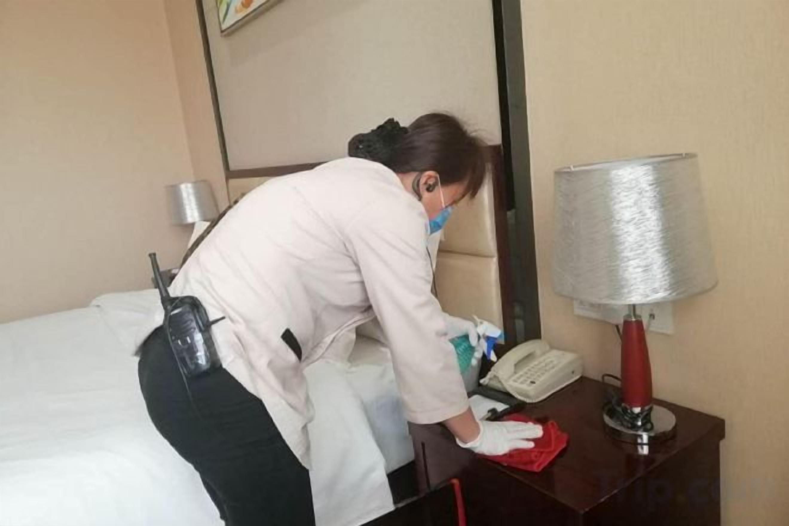 A hotel room with a bed, a bedside table with a lamp, a telephone, and a person cleaning.