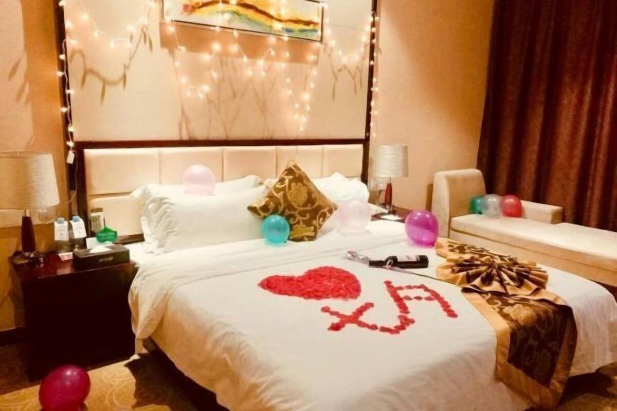 A hotel room with a bed, bedside tables, and a nightstand. There are decorative lights, a painting on the wall, and a bedspread with a heart design.