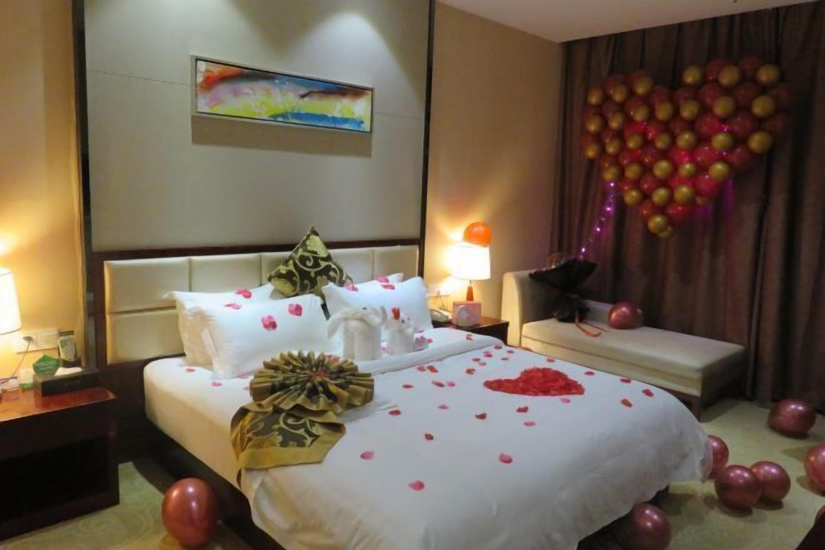 A hotel room with a bed adorned with rose petals and a heart-shaped decoration, a bedside table with a lamp, a wall-mounted painting, and a curtain with a heart-shaped balloon arrangement.