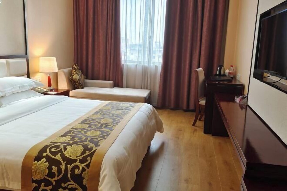 A hotel room with a large bed, a desk, a chair, a TV, and a window with curtains.