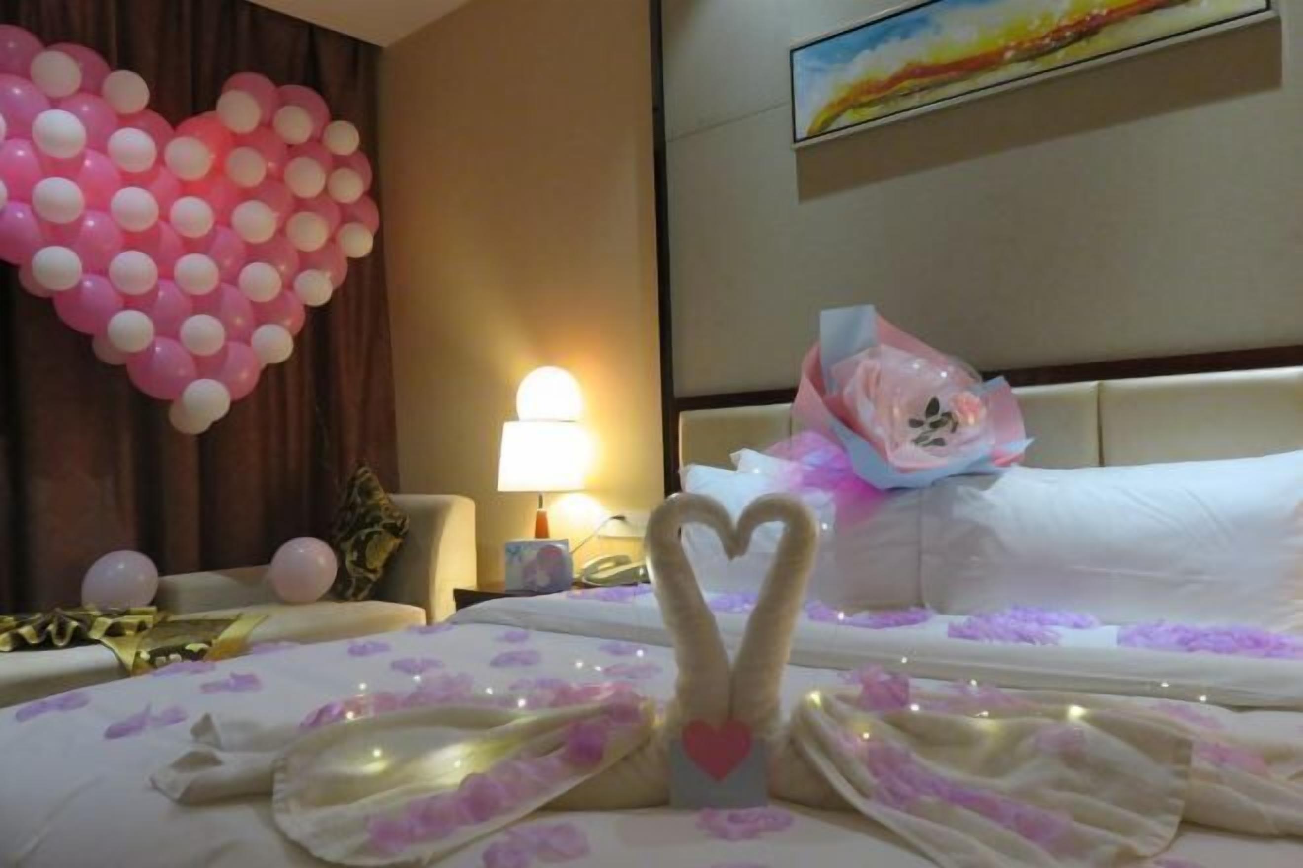 Habitación de hotel con un globo decorativo con forma de corazón, un ramo de flores y una toalla doblada en forma de cisne sobre la cama.