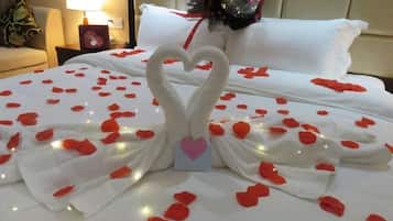Una cama con una toalla dispuesta en forma de corazón y pétalos de rosa, una lámpara de noche y una almohada decorativa.
