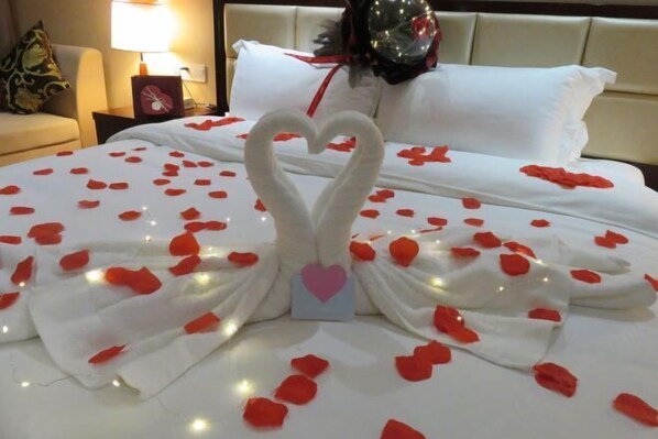A bed with a heart-shaped towel arrangement and rose petals, a bedside lamp, and a decorative pillow.