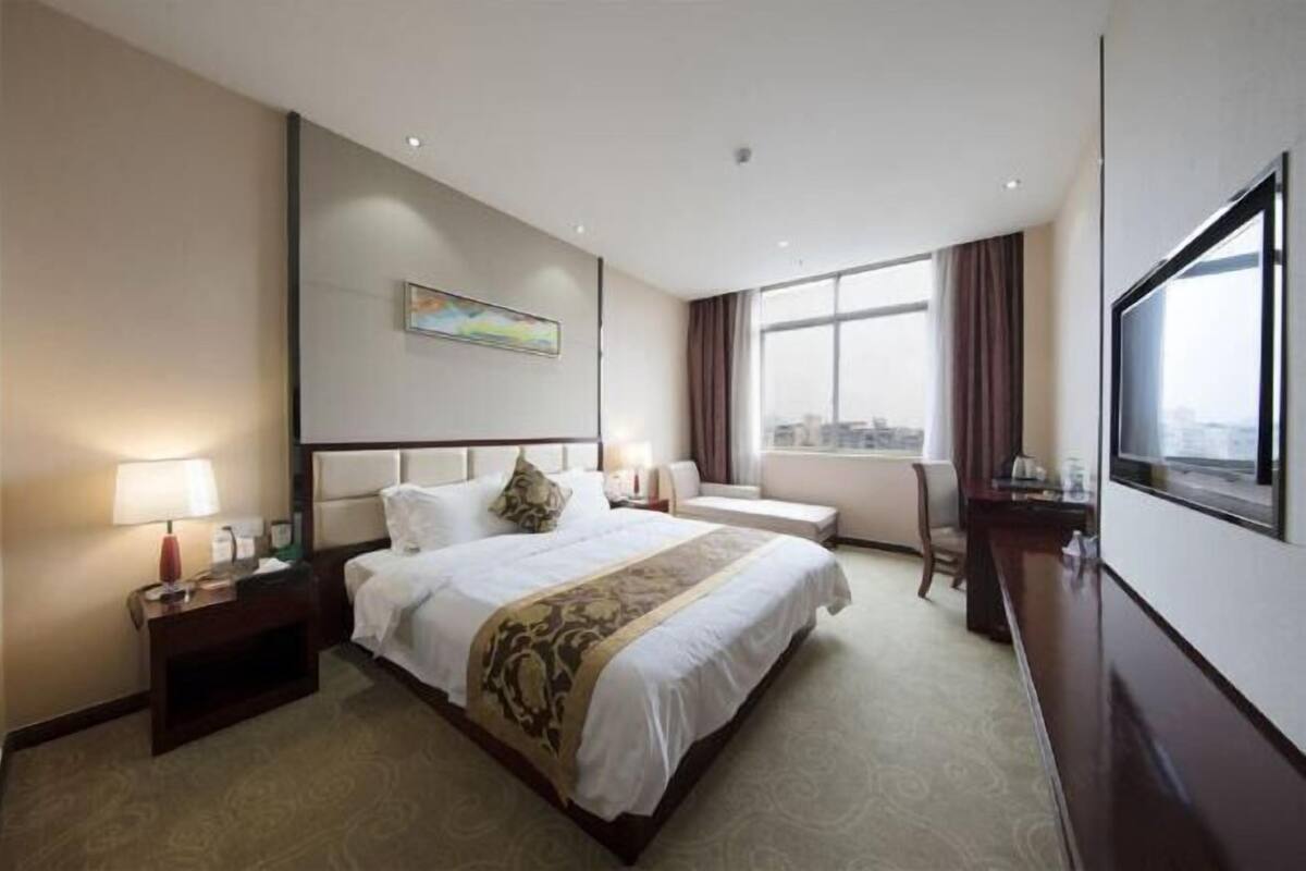 A hotel room with a large bed, a desk, a chair, a window with a city view, and a painting on the wall.