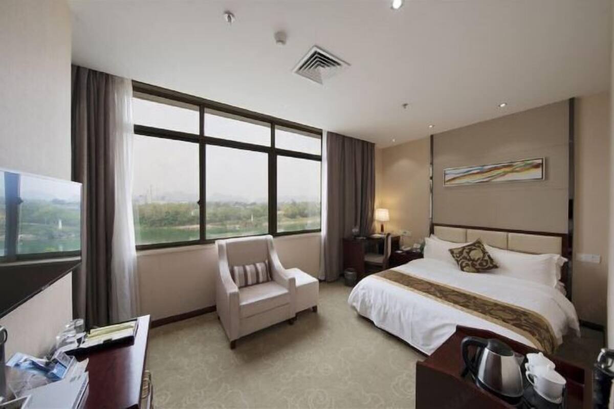 A hotel room with a large window, a bed, a chair, a desk with a computer, and a coffee maker.