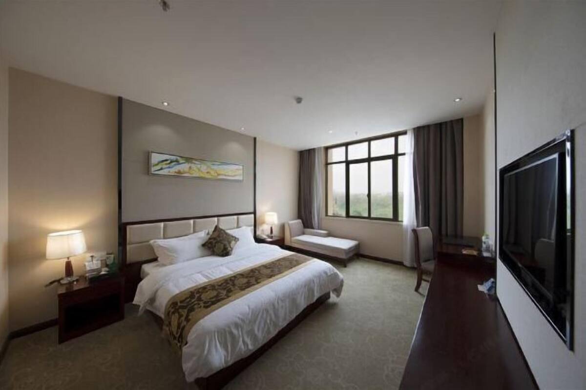 A hotel room with a large bed, bedside tables, a TV, a window with curtains, and a painting on the wall.
