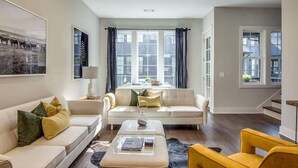 Un salon moderne avec un canapé, un pouf et un fauteuil jaune. On y trouve une fenêtre avec des rideaux, une lampe et un tableau encadré au mur.