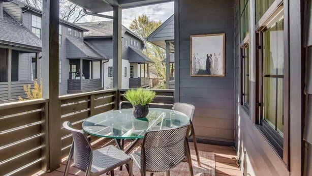 Une terrasse extérieure couverte, dotée d’une table en verre et de chaises en métal, entourée de rampes en bois et offrant une vue sur les maisons voisines.