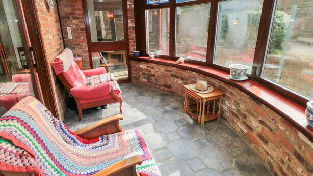 A sunroom with brick walls, large windows, a wooden bench, a red chair, and a crocheted blanket. - Eric House (Filey)