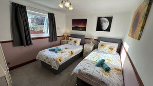 A bedroom with two beds, a window with curtains, a nightstand with a lamp, and wall art including a moon and sunflowers. - Eric House (Filey)