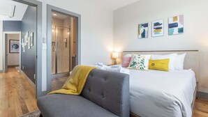 A modern hotel room with a grey sofa, a bed with white bedding and yellow pillows, a shower area, and abstract wall art.