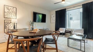 Un salon moderne avec une table de salle à manger ronde en bois, des chaises noires, une table basse en verre et un téléviseur à écran plat fixé au mur.