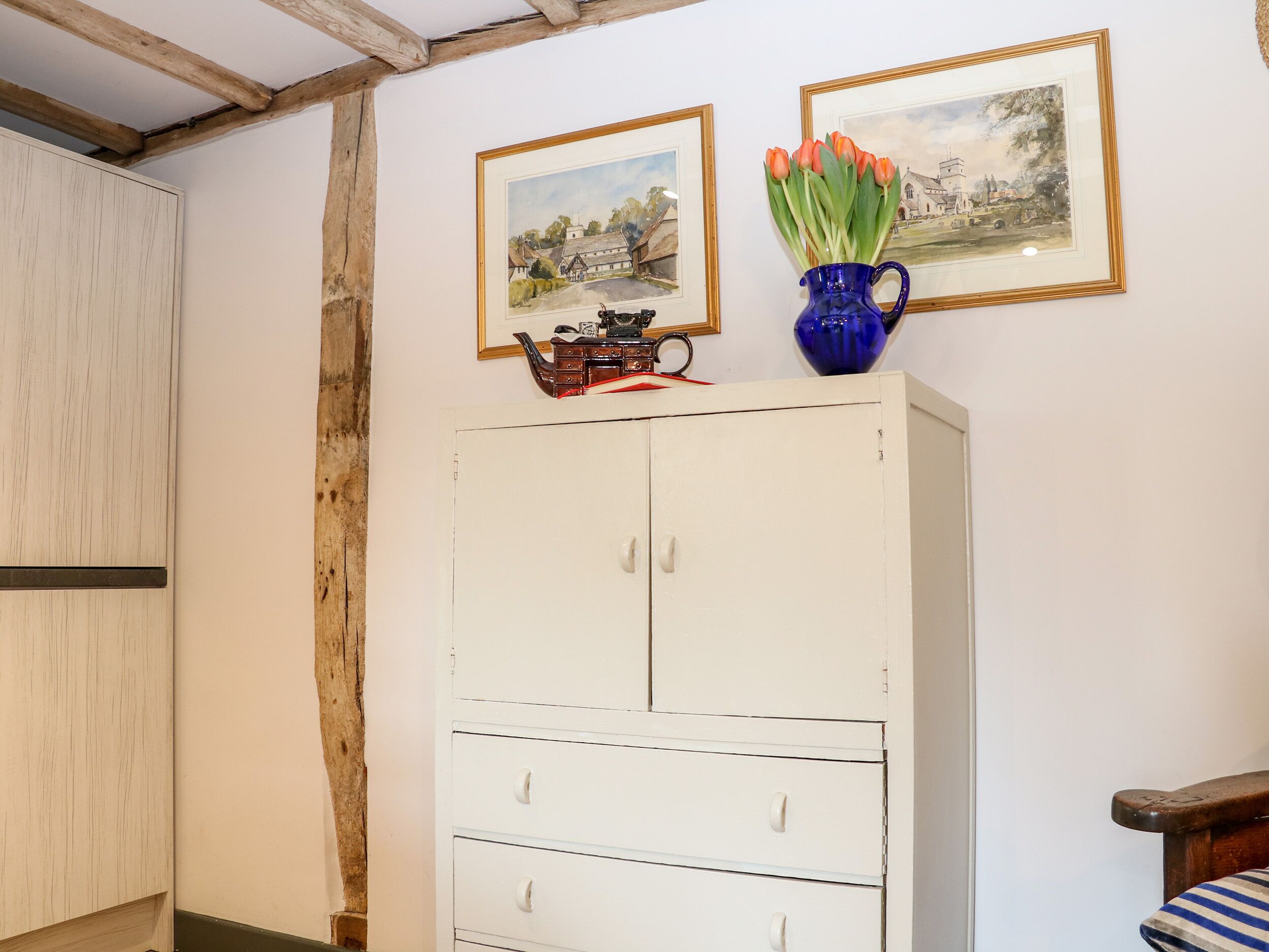 A room with a white cabinet, wooden beams on the ceiling, and two framed paintings above it.