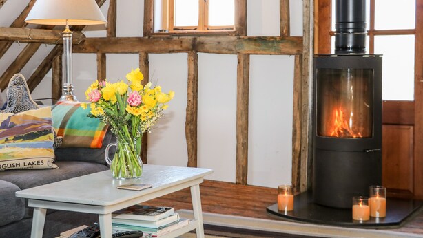 A cozy room with exposed wooden beams, a fireplace, a table with a vase of flowers, and a sofa with cushions.