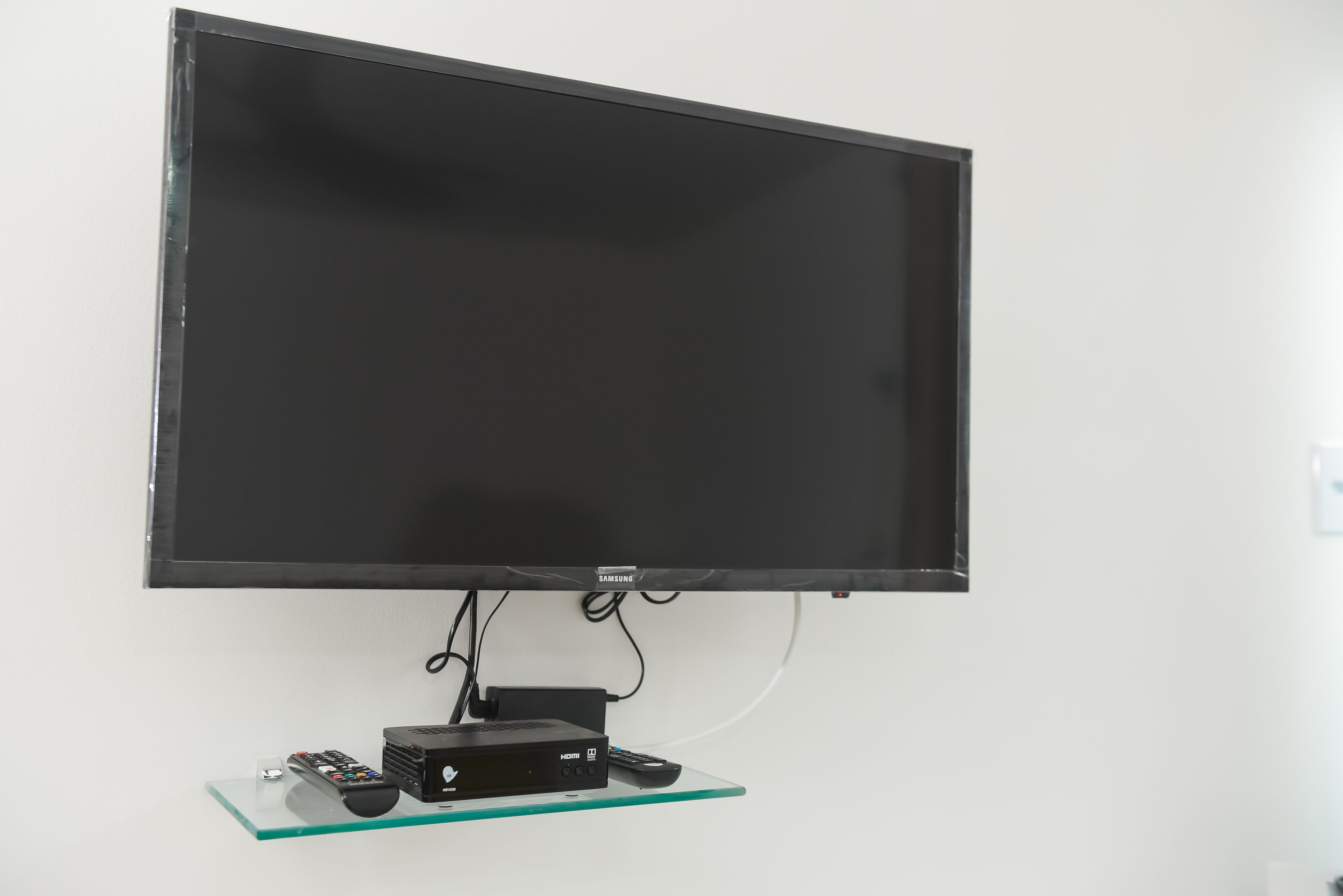 A flat-screen TV mounted on a wall with a glass shelf below it, holding a remote control and a small electronic device.
