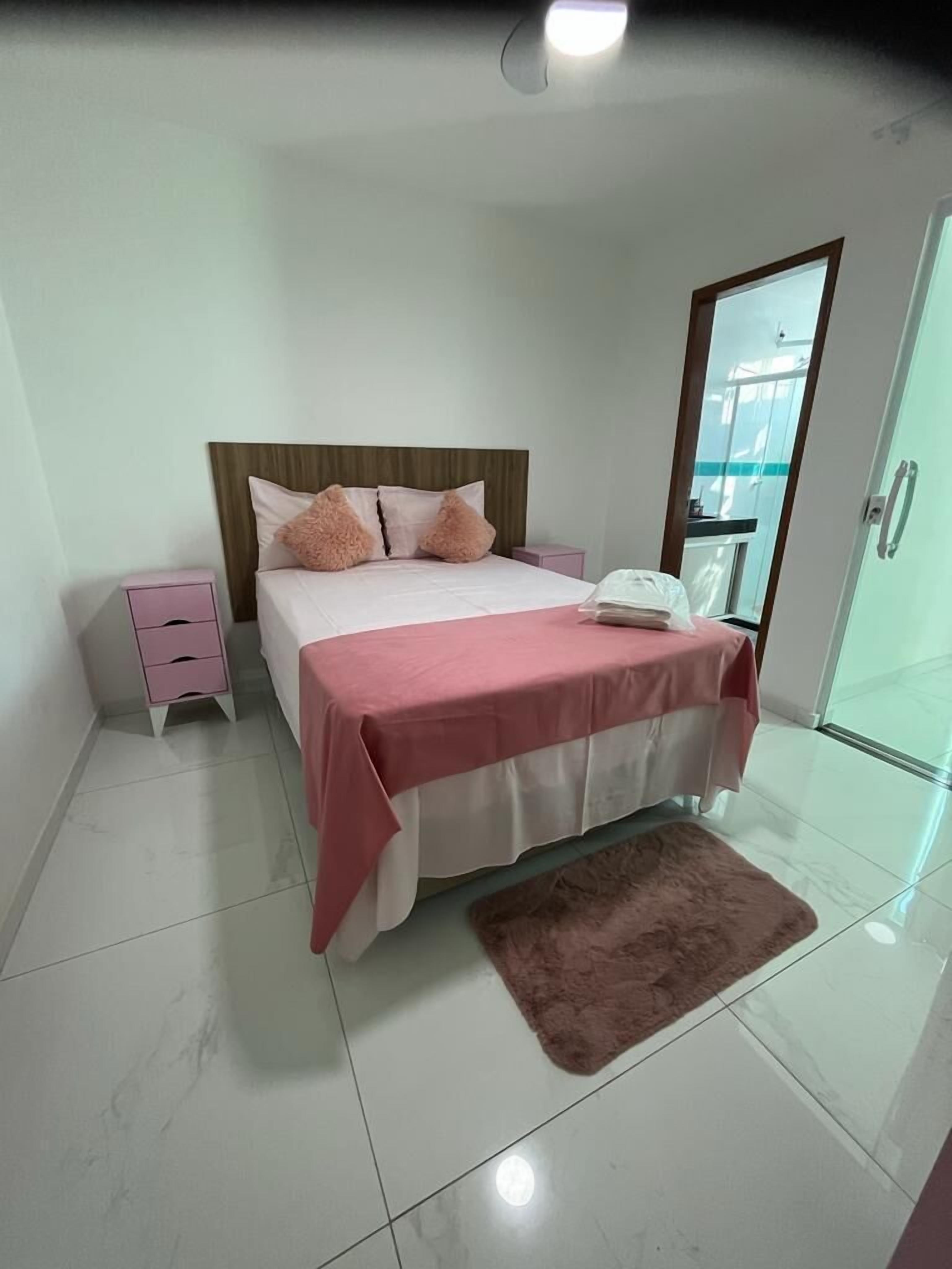 A bedroom with a bed, a bedside table, a nightstand, a bathroom door, and a bathroom mirror.