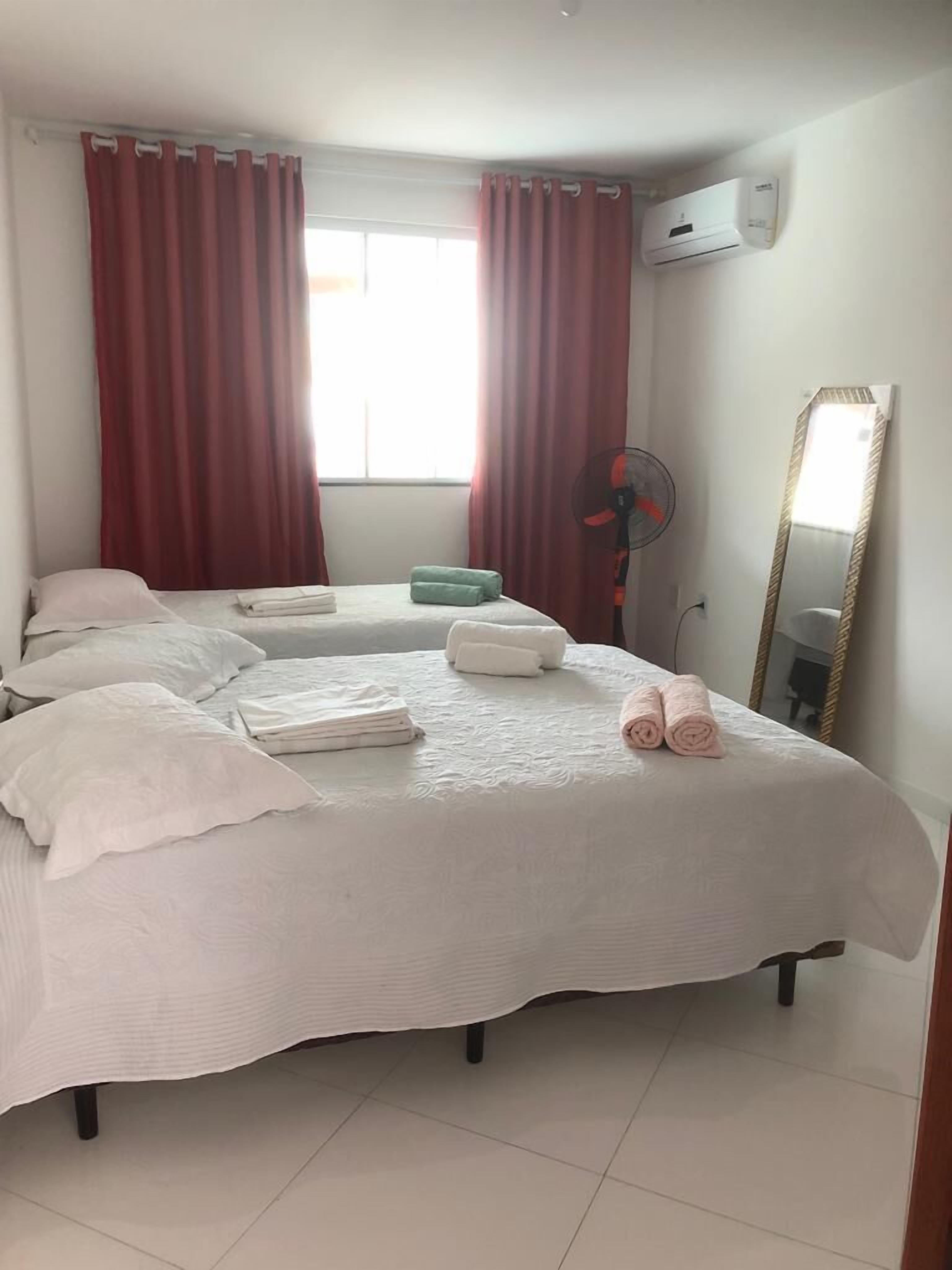 Una cama bien hecha con sábanas blancas y toallas rosas, un ventilador de pie y un espejo grande en la pared.