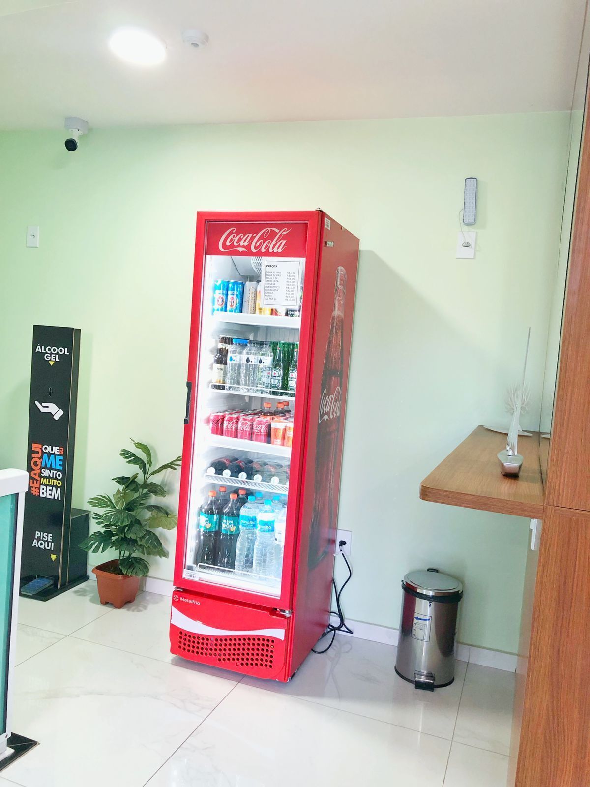 Una máquina expendedora roja de Coca-Cola repleta de diversas bebidas, una planta en maceta y un cubo de basura en una habitación con paredes verde claro y una estantería de madera.