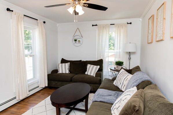 A living room with a brown sofa, a round coffee table, and a ceiling fan. There is a window with white curtains and a wall-mounted shelf with two small plants.