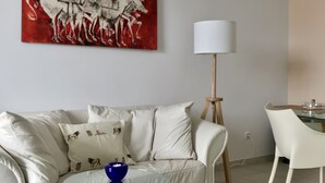 Un salon moderne avec un canapé blanc, une table basse en verre, un tableau abstrait rouge, une lampe blanche et un coin repas avec une table en verre et des chaises blanches.