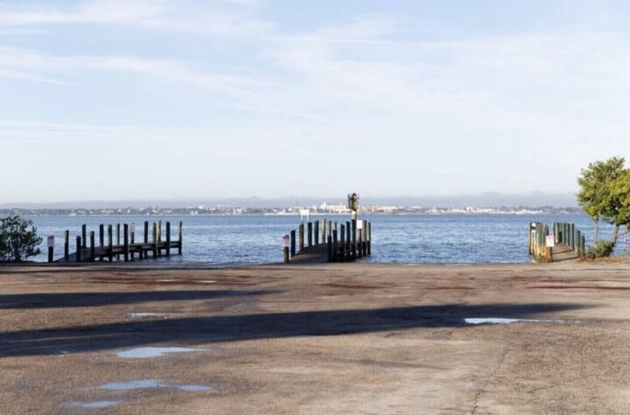 Blick auf das Wasser mit einem Betonsteg, einem kleinen Gebäude in der Ferne und blauem Himmel.