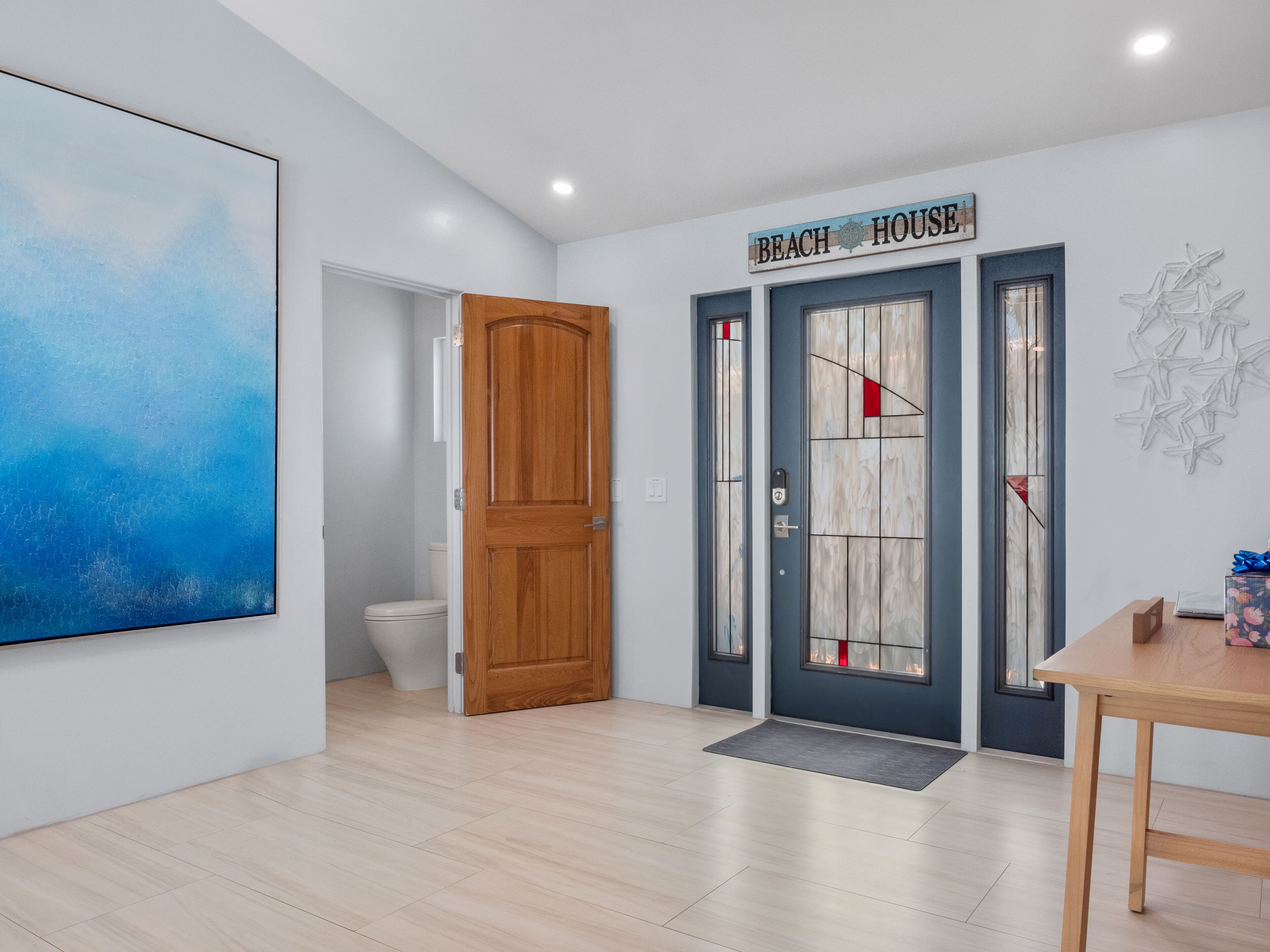 Un interior moderno con una puerta de madera, un baño, una zona de comedor y un cartel en la pared que dice 'BEACH HOUSE'.