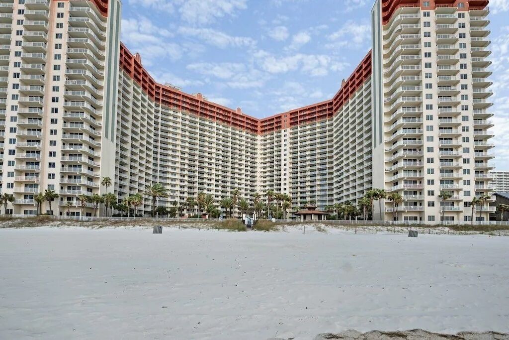 A high-rise hotel with multiple balconies, a beach view, and a sandy foreground.