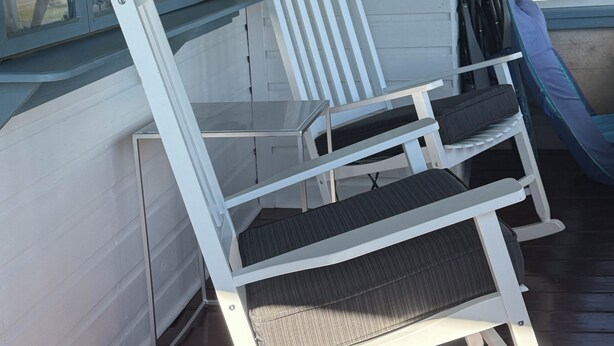 Une chaise berçante et une chaise placées devant une maison, avec une pancarte indiquant : « BIENVENUE DANS NOTRE MAISON AU BORD DU LAC, INSTALLEZ-VOUS COMME CHEZ VOUS. »