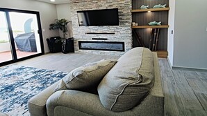 Un salon moderne comprenant un canapé, un téléviseur à écran plat fixé sur un mur en pierre, une étagère en bois ornée d’objets décoratifs et une grande fenêtre donnant sur l’extérieur.