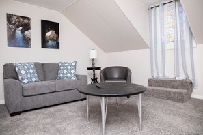 Een woonkamer met een grijze bank, een zwarte tafel en een stoel. Twee ingelijste foto's hangen aan de muur, er staat een lamp en er is een raam met gordijnen.