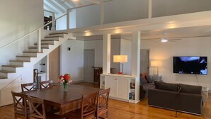 A spacious living area with a wooden dining table, chairs, a sofa, a TV, and a staircase.