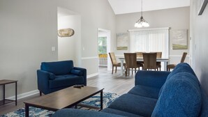 Una sala de estar con un sofá azul, un sillón azul, una mesa de centro de madera y un comedor con mesa y sillas.