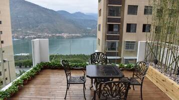 A balcony with a wooden deck, metal chairs, and a table. A mountain and a body of water are visible in the background.