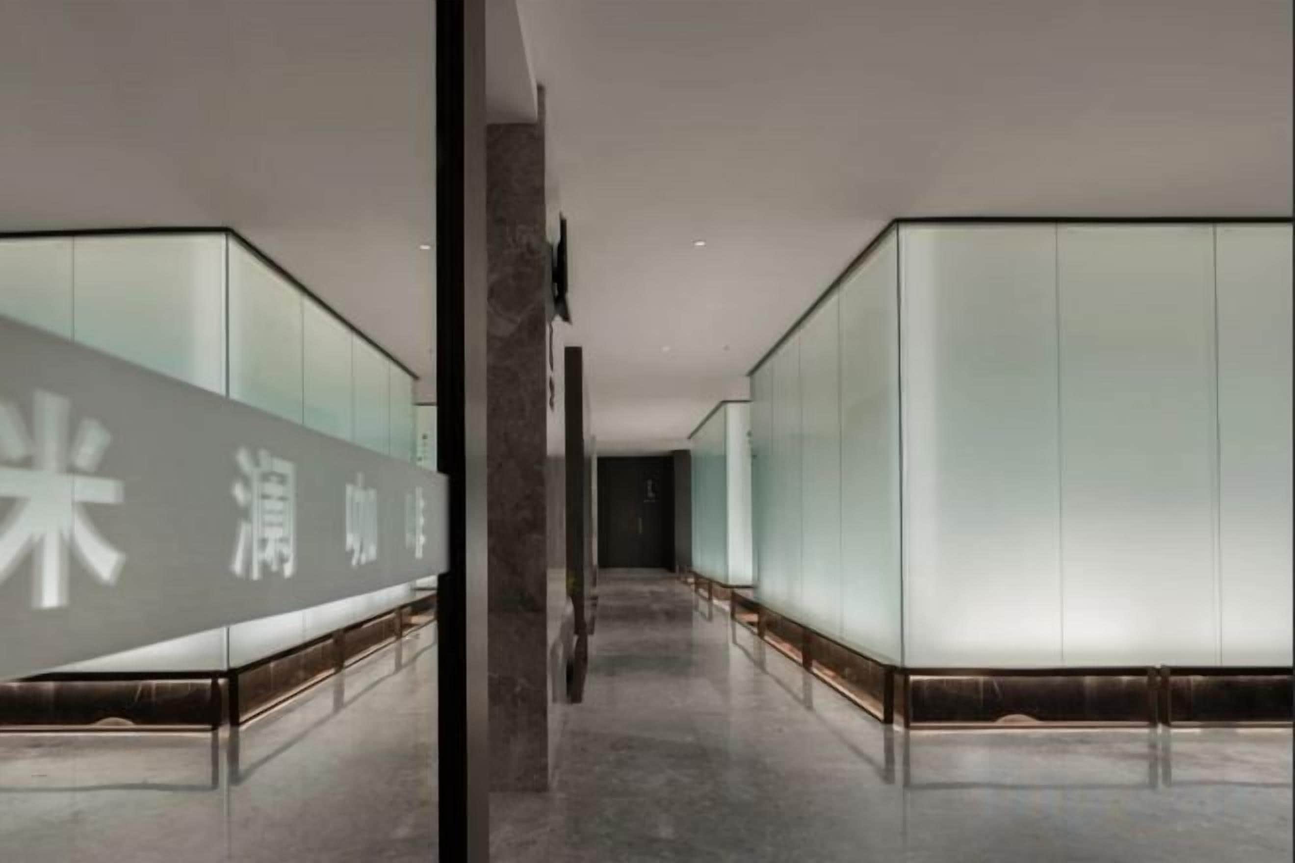 A corridor with mirrored walls and a reflective floor, leading to a room with a large mirror and a door.