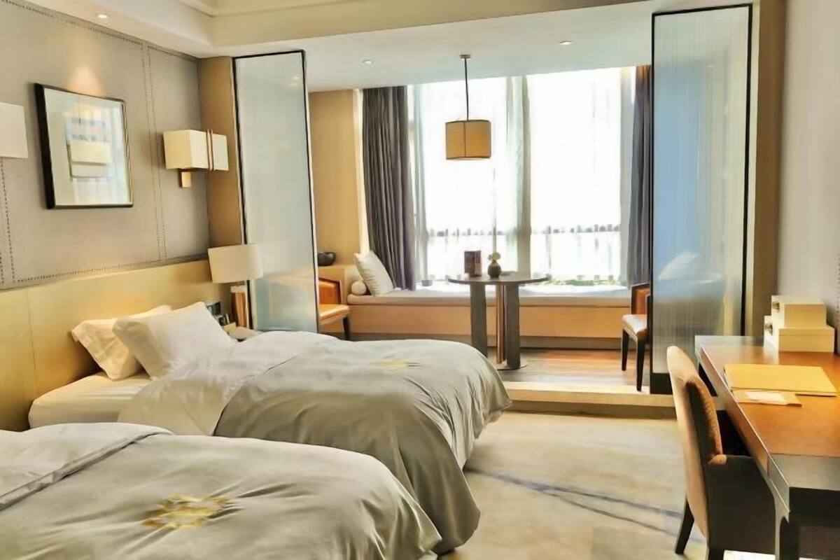 Habitación de hotel con dos camas, un escritorio, una silla, un ventanal con cortinas y una mesita con lámpara.