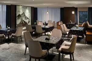 A hotel dining area with multiple tables and chairs, a large painting on the wall, and a view of the outdoors through the windows.