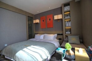 Habitación de hotel con una cama grande, una mesita de noche con lámpara, una pequeña zona de estar con cojín y una estantería con libros y objetos decorativos.
