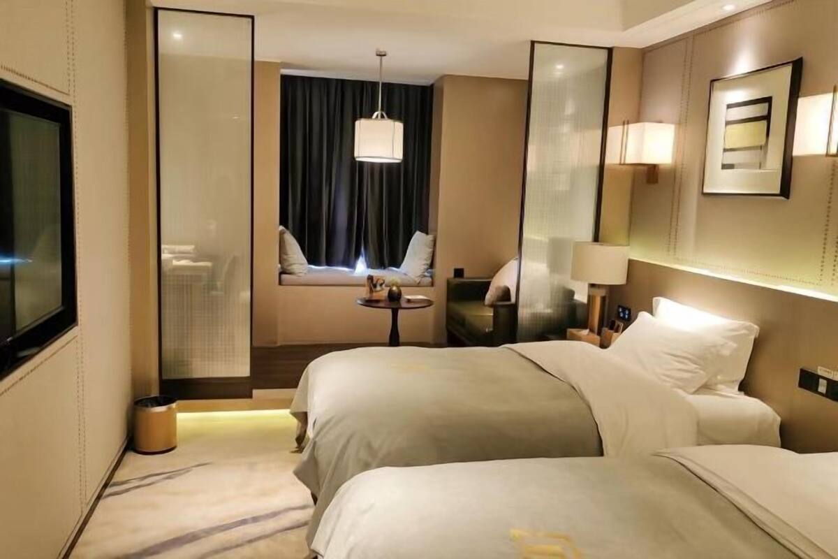 Habitación de hotel con dos camas, una mesita, una silla, un ventanal con cortinas, un espejo y una lámpara de pared.