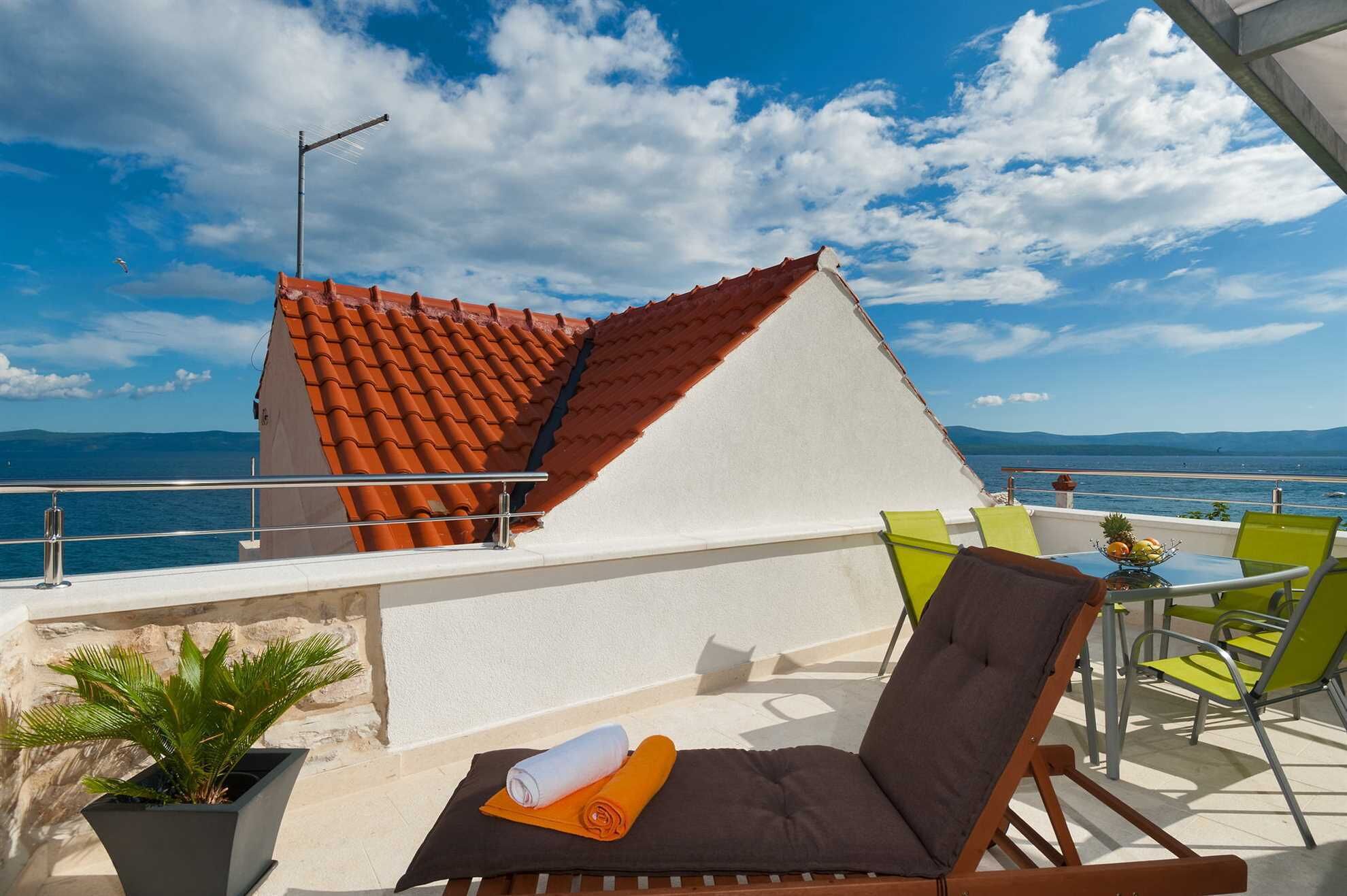 Une terrasse sur le toit, avec un toit recouvert de tuiles rouges, un mur blanc et une vue sur la mer. On y trouve des chaises vertes et jaunes, une petite table avec des fruits et une plante en pot.