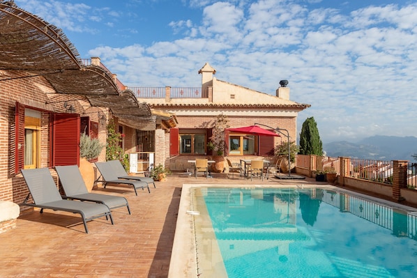 A poolside area with lounge chairs, a pool, and a building with a red roof and brick walls.